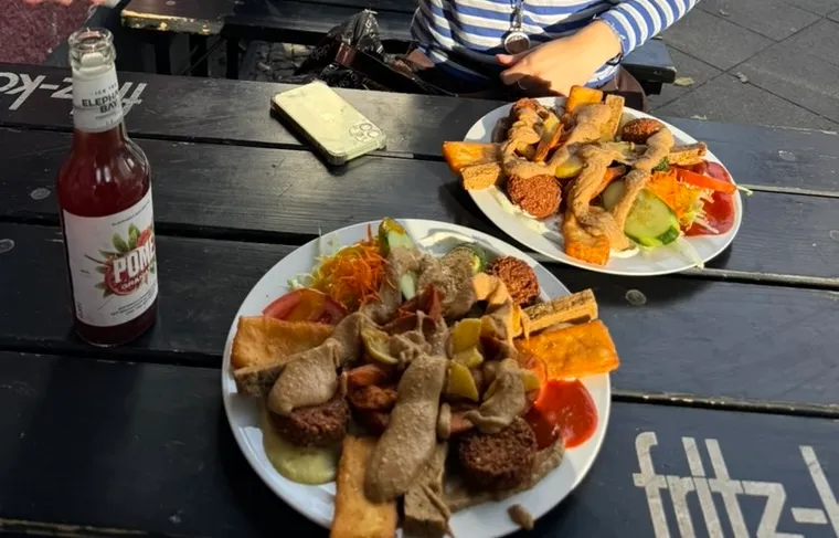 Zwei volle Teller mit Falafel, Tofu, Halloumi, Salat und Soße auf einem schwarzen Biertisch, daneben eine Limo-Flasche